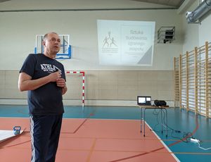 hala sportowa, po lewej Paweł Ochwat, po prawej ekran ze slajdem wyświetlanym na rzutniku, widoczny tytuł slajdu: Sztuka budowania zgranego zespołu.