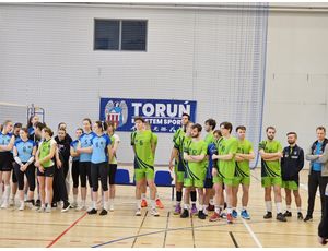 Grupa osób w strojach sportowych, stojących wokół boiska do siatkówki, gotowych do rozegrania meczu.